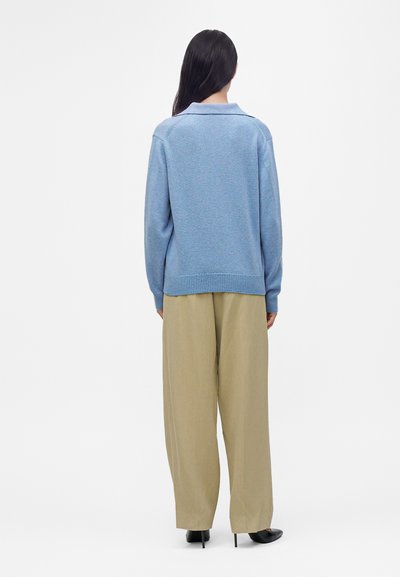 Femme aux longs cheveux foncés portant un pull bleu clair, un pantalon large beige et des chaussures à talons noirs, vue de dos.