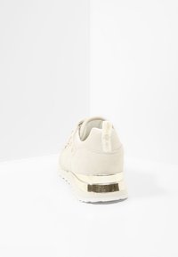 Baskets en suède beige clair avec un talon doré métallique, design lacé et petit détail de logo à l'arrière. Style texturé et moderne.