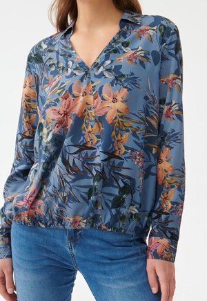 Femme portant un chemisier bleu à manches longues avec un motif floral coloré et un ourlet élastique, associé à un jean bleu.