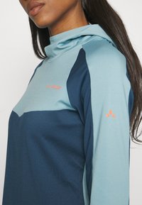 Blå och teal huvtröja med hög krage. Har kontrasterande paneler, subtil textur och en orange varumärkeslogotyp på bröstet och ärmen.