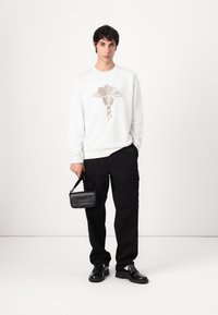 El sudadera blanca presenta un diseño geométrico en beige, combinado con pantalones cargo negros y un pequeño bolso negro. Lleva zapatos negros.