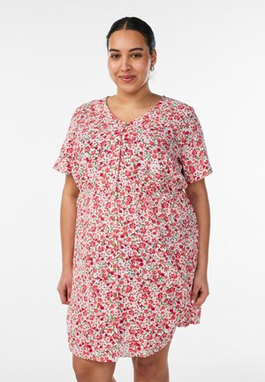 Kvinde med mørkt hår trukket tilbage iført en kortærmet, knælængde kjole med et rødt og lyserødt blomsterprint, stående mod en hvid baggrund.