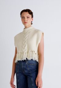 Strikket ermeløs genser i kremfarge med kabelmønster, høy hals og frynset nederkant. Kombinert med høyt liv mørke denimjeans.