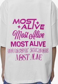 Hvit overdimensjonert t-skjorte med dristig, rosa tekst som viser forskjellige stiler av «Most Alive» og «Most Alive» i ulike skrifttyper og størrelser.
