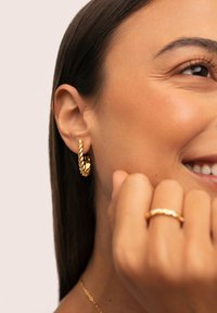 Orecchini a cerchio color oro con un design intrecciato, caratterizzati da accenti testurizzati, indossati da una modella. Un anello intrecciato coordinato è sulla sua mano.