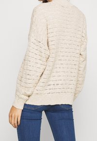 Beige stickad tröja med en lös passform, ribbade ärmslut och ett strukturerat mönster. Har neddragna axlar och en hög halsringning.