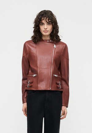 Jonge vrouw met krullend haar, gekleed in een nauwsluitend bruine leren jas met een asymmetrische ritssluiting en gespen, gecombineerd met een zwarte broek.