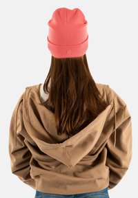 Persona con lunghi capelli castani, che indossa un berretto rosa, una felpa beige e jeans blu, vista da dietro su uno sfondo bianco.