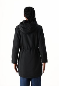 Svart hetteparka med snøring i midjen, som rekker til midt på låret. Har lange ermer og myk tekstilfôr inne i hetten.