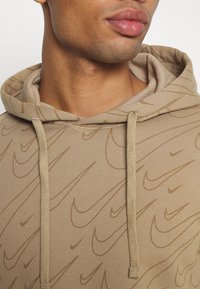 Beige hoodie med ett subtilt tonalt Nike-logomönster, utformad med en dragsko och textilstruktur. Närbild som betonar halslinningen.