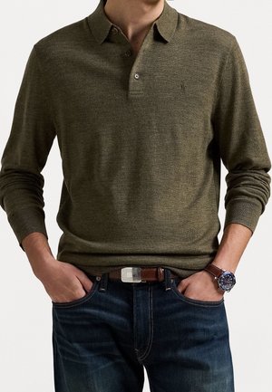 Mann trägt olivgrünes langärmeliges Polohemd, dunkelblaue Jeans, braunen Gürtel und dunkle Uhr mit Lederarmband, Hände in den Taschen.