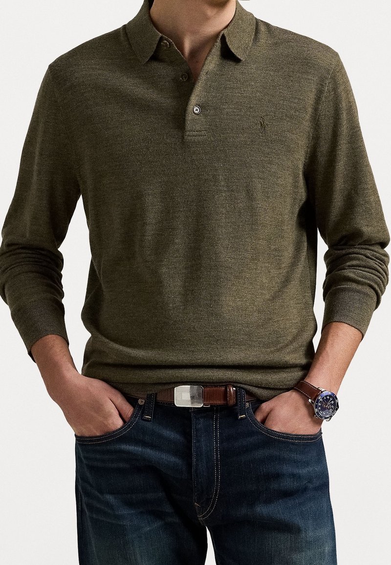 Homme portant un polo manches longues vert olive, jean bleu foncé, ceinture marron et montre sombre avec bracelet en cuir, mains dans les poches.