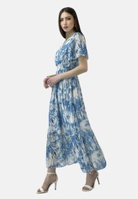 Robe maxi florale bleu et blanche ; tissu léger avec des manches courtes, taille smockée et jupe fluide. Assortie à des talons blancs.