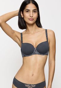 Nainen, jolla on päällään tummanharmaat yhteensopivat rintaliivit ja alushousut, joissa on pitsiyksityiskohtia, poseeraa tasaisella valkoisella taustalla.