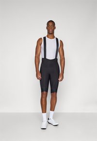 Schwarze Radsport-Bib-Shorts mit weißem Mesh-Unterhemd, ausgestattet mit schwarzen Trägern, knielangem Schnitt und glatter Textur. Weiße Sportschuhe und Socken.