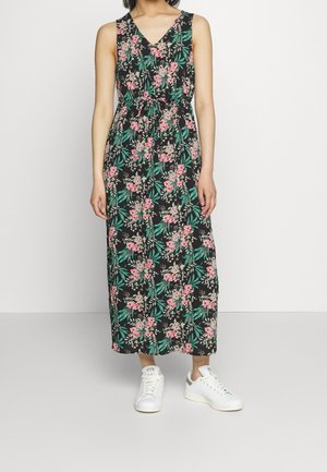 Kvinde iført en sort blomster maxi kjole uden ærmer med grønne blade og pink blomster, kombineret med hvide sneakers på ensfarvet baggrund.