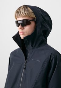 Black waterproof jacket with a high collar and zipper. Worn with dark, angular sunglasses. Smooth texture and no visible patterns.