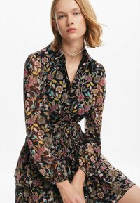 Blommig klänning med svart genomskinligt tyg, krage, långa ärmar och åtsittande midja. Har volangdetaljer och färgglada mönster.