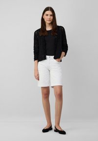 Zwarte kanten cardigan, aansluitend zwart topje, witte denim shorts en zwarte flats. De cardigan heeft een knoopsluiting aan de voorkant en een open geweven ontwerp.