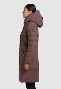 Barna puffkabát magas gallérral és kapucnival, gyémánt alakú steppeléssel, rugalmas oldalsó panellel és oldalsó zsebekkel.