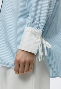 Χέρι που φοράει μπλε και λευκό ριγέ πουκάμισο με μακριά μανίκια, φαρδύ λευκό μανσέτι και λεπτομέρεια με κορδέλα δεμένη, σε συνδυασμό με λευκό παντελόνι.