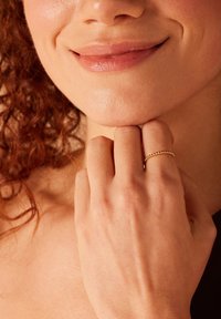 Bague en or avec un motif perlé, portée à l'index, mettant en valeur une peau lisse et des textures naturelles autour de la main et du visage.