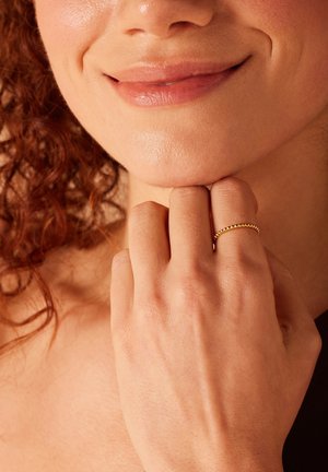 Gouden ring met een kralenpatroon, gedragen op de wijsvinger, die een gladde huid en natuurlijke texturen rond de hand en het gezicht toont.