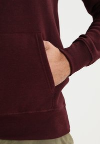 Persona che indossa un hoodie bordeaux con una mano nella tasca frontale, su uno sfondo chiaro e uniforme.