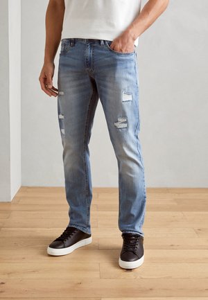 Mand iført lyseblå slim-fit slidte jeans og sorte sneakers med hvide såler, stående på trægulv.