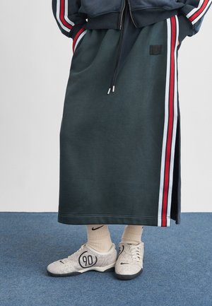 Osoba oblečená v dlouhé tmavé sukni s červenými, bílými a modrými pruhy, bílých ponožkách Nike a bílých teniskách s potiskem "90".