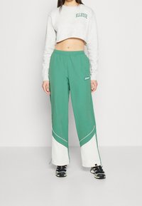 Felpa grigia tagliata con testo "ELLESSE", abbinata a pantaloni larghi verdi con accenti bianchi e logo sulla coscia. Sneakers nere.