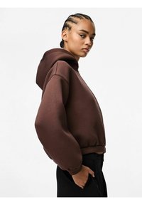 Felpa marrone con cappuccio, dal taglio morbido, con polsini e orlo a costine. Tessuto liscio con tasca a marsupio, indossata con pantaloni neri.