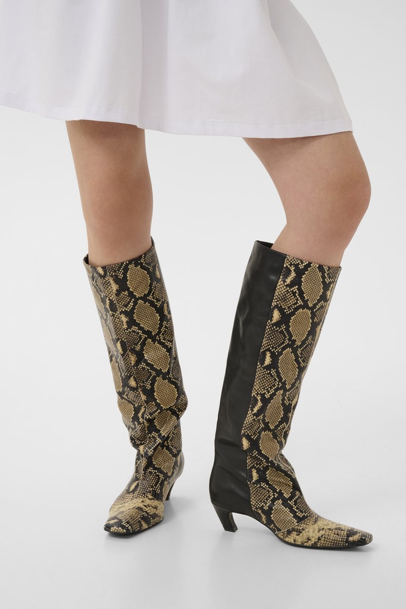 Femme portant des bottes montantes à imprimé serpent avec des panneaux en cuir noir, assorties à une jupe blanche.