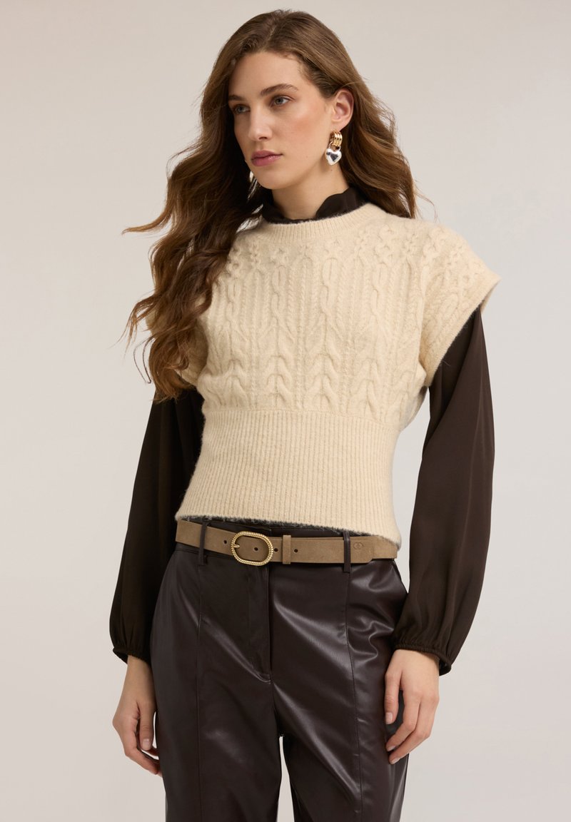 Kremowy bezrękawnik z warkoczowym splotem i ściągaczem u dołu, noszony na ciemnej koszuli. Połączony z wysokim stanem błyszczących brązowych spodni ze sztucznej skóry i beżowym paskiem.