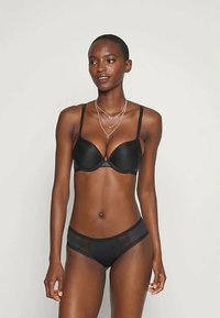 Žena v čiernej podprsenke a zodpovedajúcom čiernom jemnom prádle, stojaca na svetlom pozadí, mírne sa pozerajúca doprava.