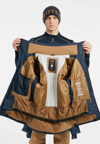 Chaqueta impermeable en color azul marino y beige con un forro interno acolchado, puños ajustables y múltiples bolsillos interiores. Presenta una etiqueta de logo cerca del cuello.
