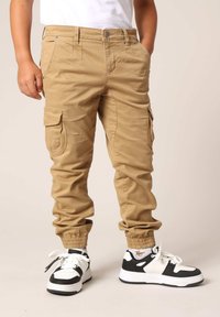 Khaki cargobroek met elastische boorden, voorzien van zijzakken en een slim fit. Gecombineerd met zwart-witte sportschoenen.