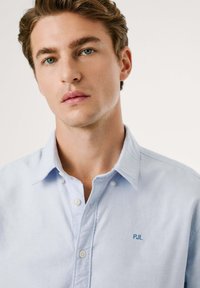 Lys blå skjorte med knapper laget av bomull, med klassisk krage, hvite knapper og en liten brodert "PJL"-logo på brystet.