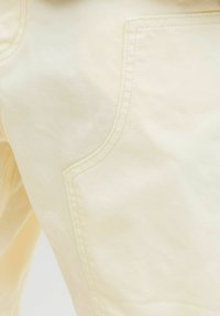 Közelkép bézs szövetből, látható varrással és nagy zsebbel, amely bemutatja a ruhadarab textúráját és varrási részleteit.