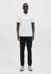 Witte poloshirt met een kraag en drie knopen, gecombineerd met donkere broeken en beige schoenen. Eenvoudig ontwerp, zachte stoftextuur.