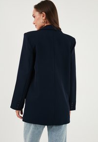 Nő, aki oversize sötétkék zakót és világoskék farmert visel, háttal áll, látható arany fülbevalókkal és hosszú barna hajjal.