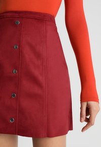 Mini gonna in suede rossa con chiusura a bottoni anteriore, cuciture verticali e un orlo leggermente svasato. Il tessuto sembra morbido e presenta una finitura opaca.