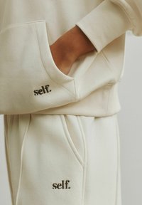 Felpa color crema con tasca a marsupio, con la scritta "self." ricamata in marrone scuro. Tessuto liscio, vestibilità comoda. Pantaloni coordinati visibili.