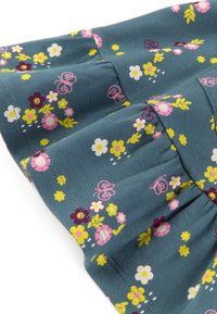 Tissu en coton bleu avec des motifs floraux en rose, jaune et blanc. Comprend un ourlet à volants et des détails de couture visibles.