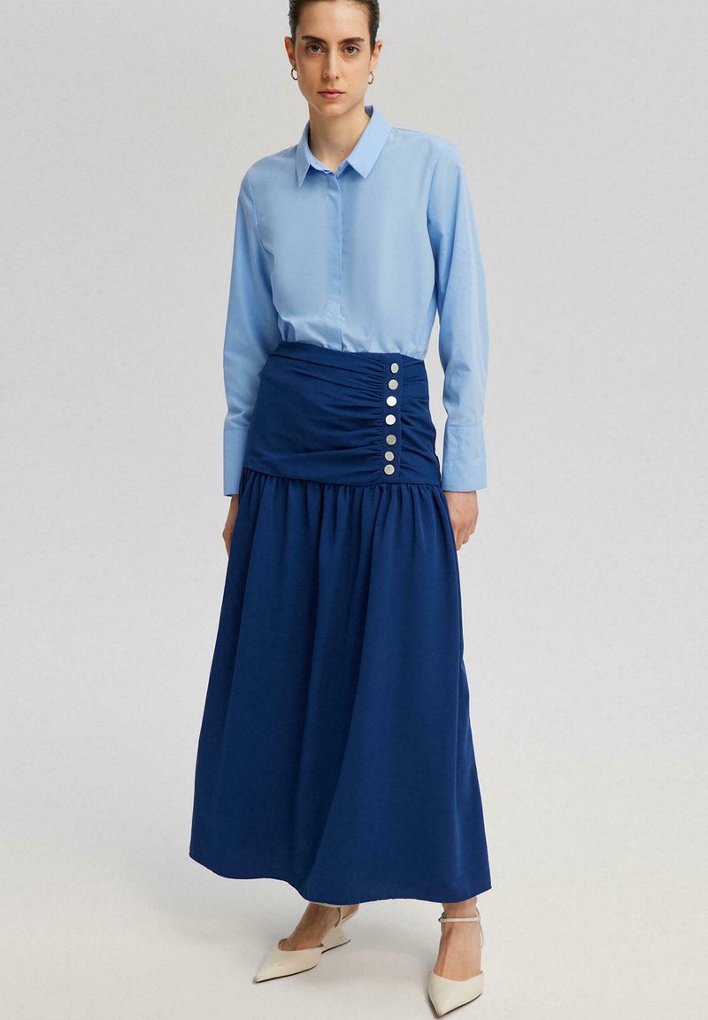 Lichtblauw langemouwenshirt gecombineerd met een marineblauwe maxi rok met ruches en zilveren knoopdetails aan de zijkant. Gladde stof met een vloeiende silhouet.