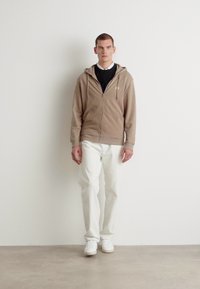 Felpa beige con zip e coulisse, indossata sopra una maglietta nera, abbinata a pantaloni bianchi e sneaker bianche, su uno sfondo neutro.