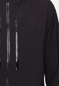 Black waterproof jacket with a smooth exterior, two zippered pockets, and a high collar; features reflective zip details.