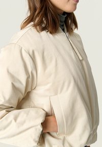 Beige bomberjack met een gladde textuur, ritssluiting, zijzakken en geribbelde manchetten. Heeft een ontspannen pasvorm en een minimalistisch ontwerp.