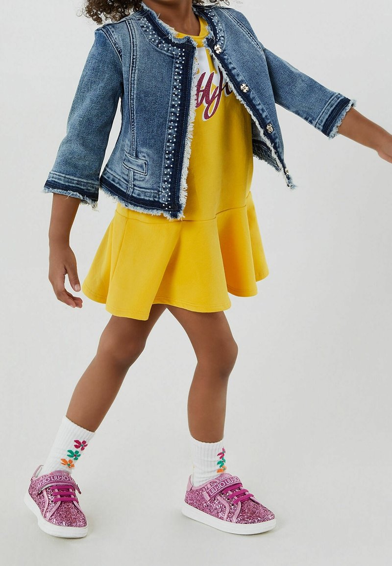 Džínová bunda s třepícími se okraji a perlovými akcenty přes žluté šaty s rozšířenou sukní. Růžové třpytivé tenisky a bílé ponožky s květinovým vzorem.