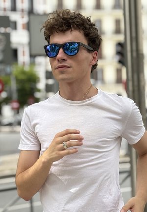 Sunglasses with dark blue lenses, black frame. Model wears a white fitted t-shirt and a silver ring, set against an urban background.